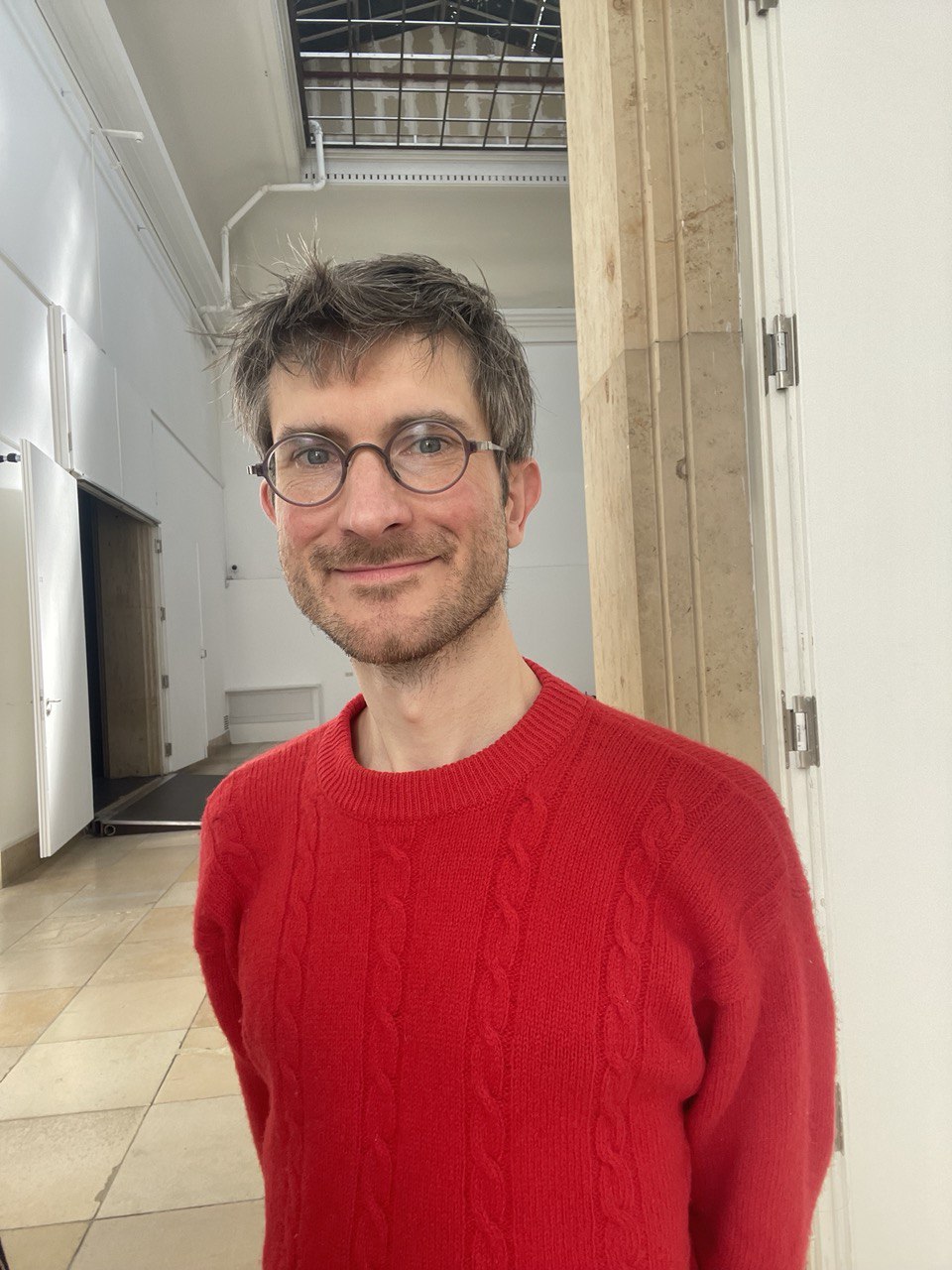 You can see Maximilian Wahlich. He is smiling, wearing round glasses and a red sweater. His hands are behind his back.