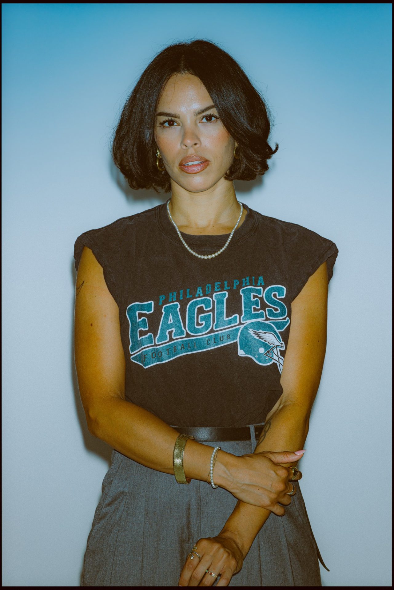 Sophie Yukiko can be seen in front of a blue background. She is wearing a brown sleeveless top with Philadelphia Eagles lettering on it. She is also wearing gray trousers. She is holding her left forearm with her right hand.