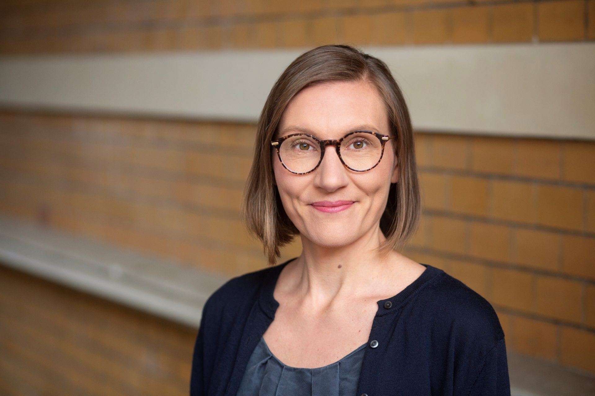 You can see Julia Schmitz's upper body. She is smiling at the camera, wearing brown glasses and a blue cardigan.