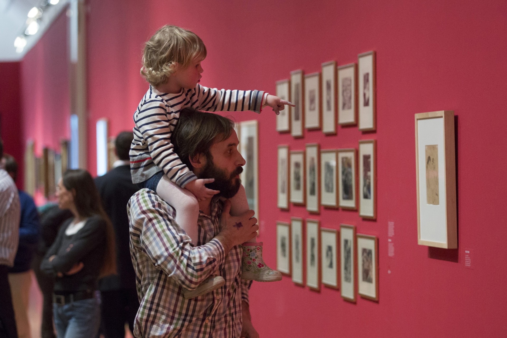 Ein kleiner Junge zeigt in einer Galerie auf Kunstwerke, während er auf den Schultern seines Vaters sitzt.