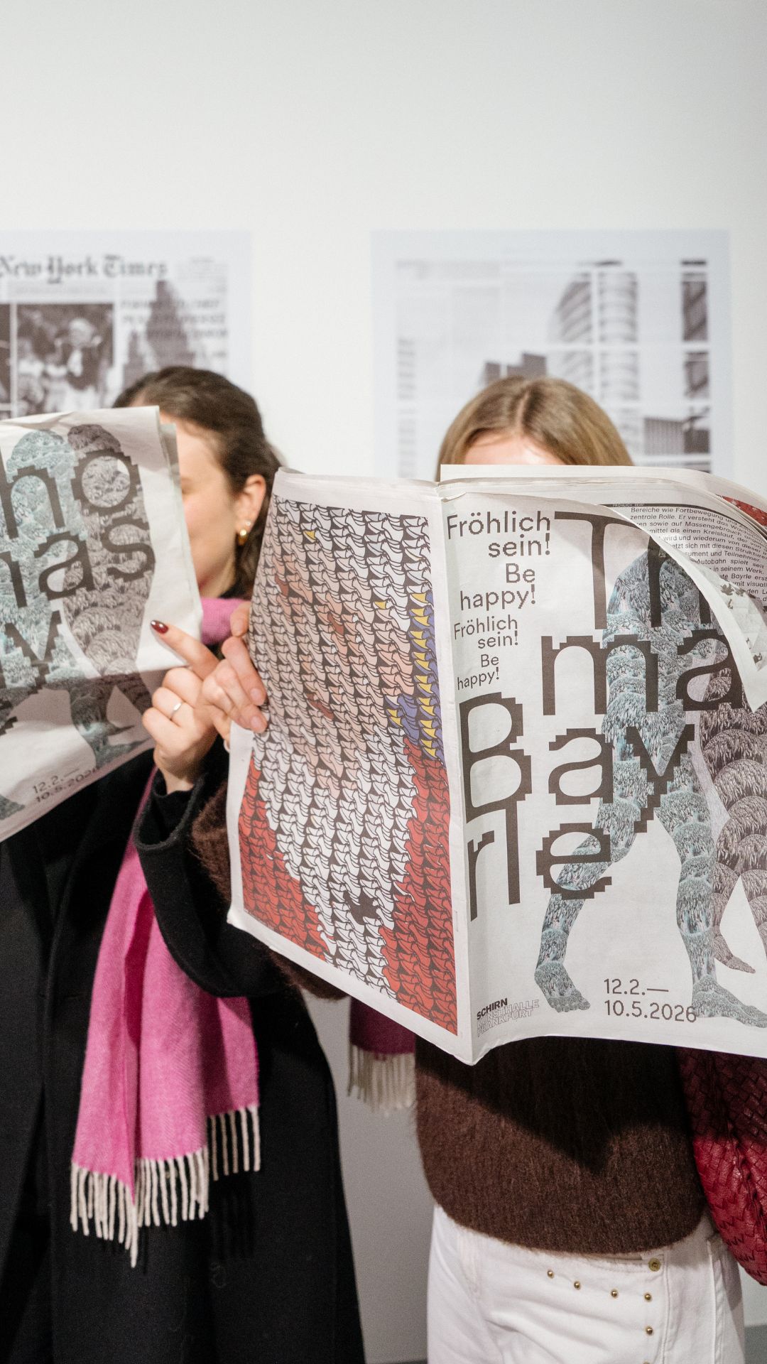 Two people hold newspapers while standing in a gallery. The newspapers feature colorful, artistic designs.