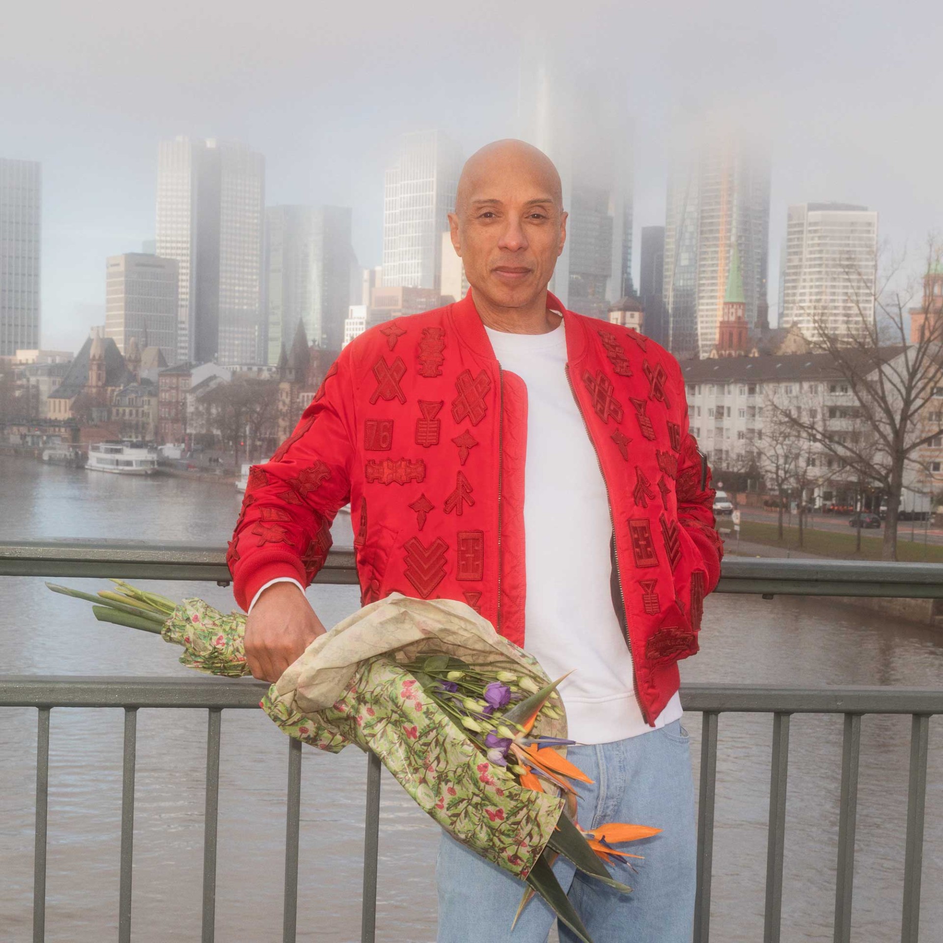 Mann in roter Bomberjacke hält einen Blumenstrauß vor einer nebligen Stadtlandschaft.