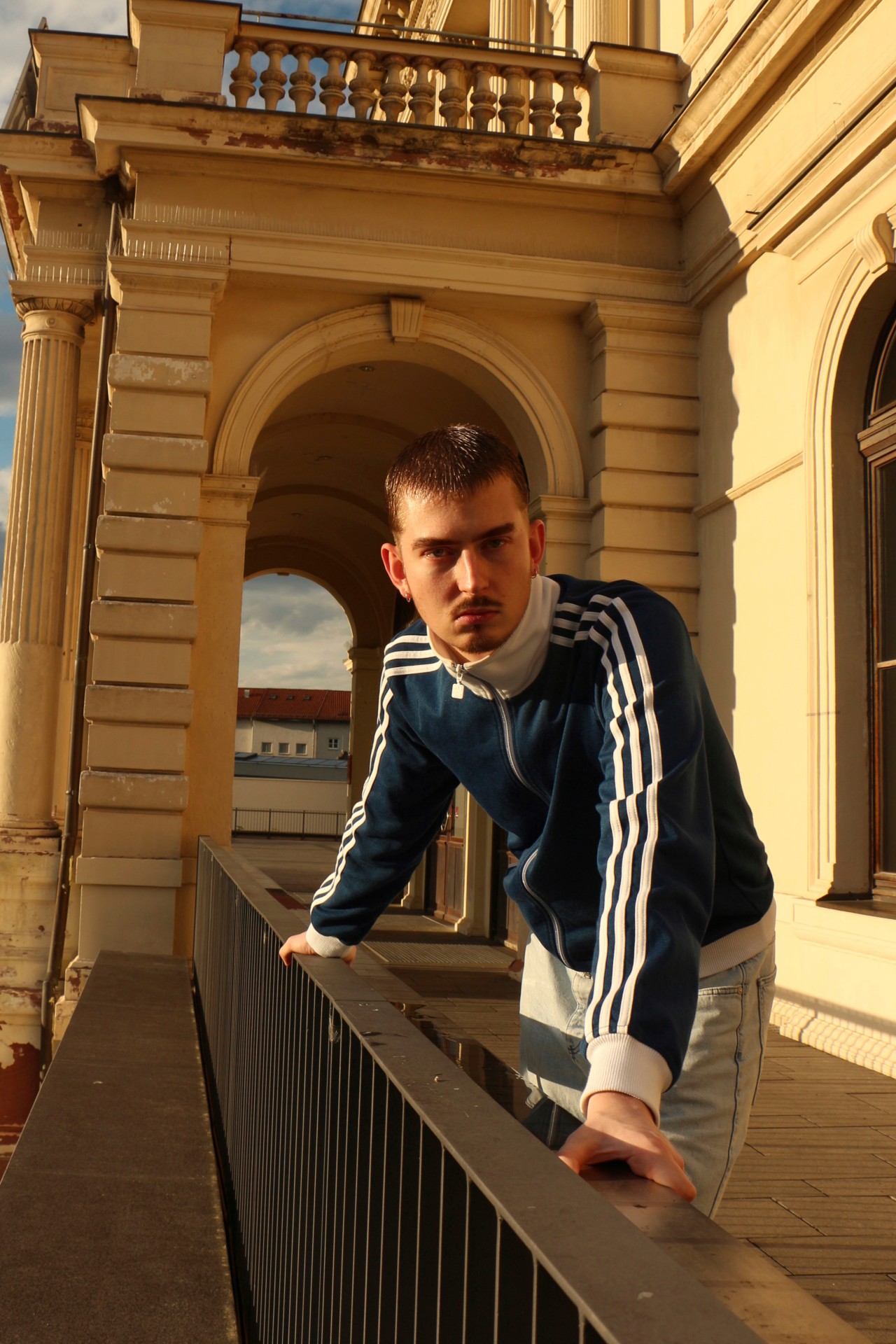 Junger Mann in blauer Sportjacke lehnt an einem Geländer auf einem Balkon mit historischer Architektur im Hintergrund.