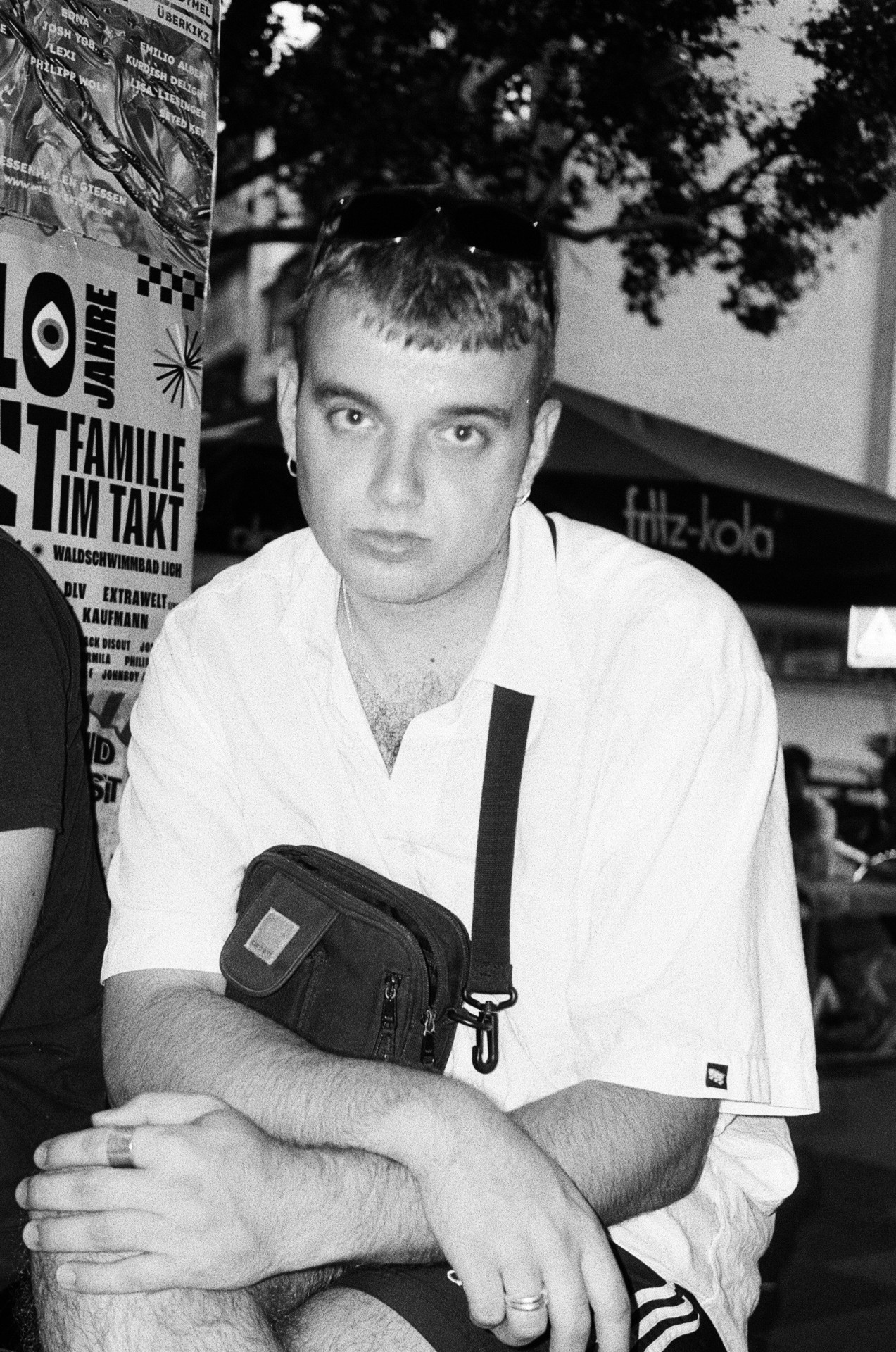 Junger Mann in Schwarz-Weiß, mit kurzer Frisur und weißem Hemd, trägt eine Umhängetasche und schaut nachdenklich.