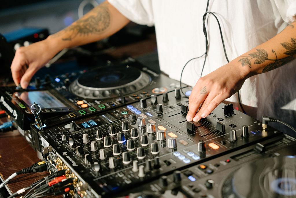 DJ mit tätowierten Händen am Mischpult, umgeben von professioneller DJ-Ausrüstung.