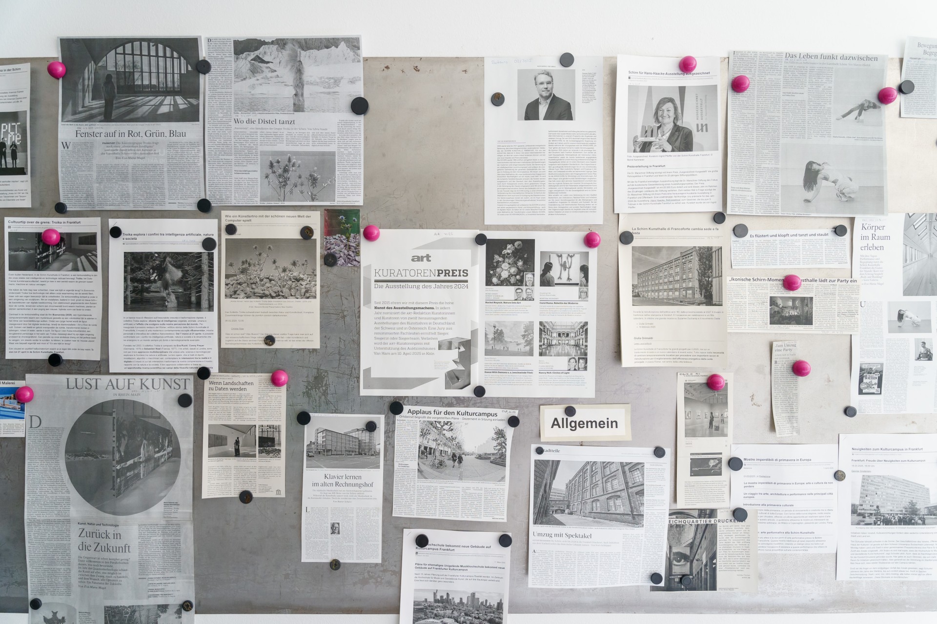 Wand mit diversen Zeitungsartikeln, Plakaten und Notizen, befestigt mit schwarzen und pinken Pinnnadeln.