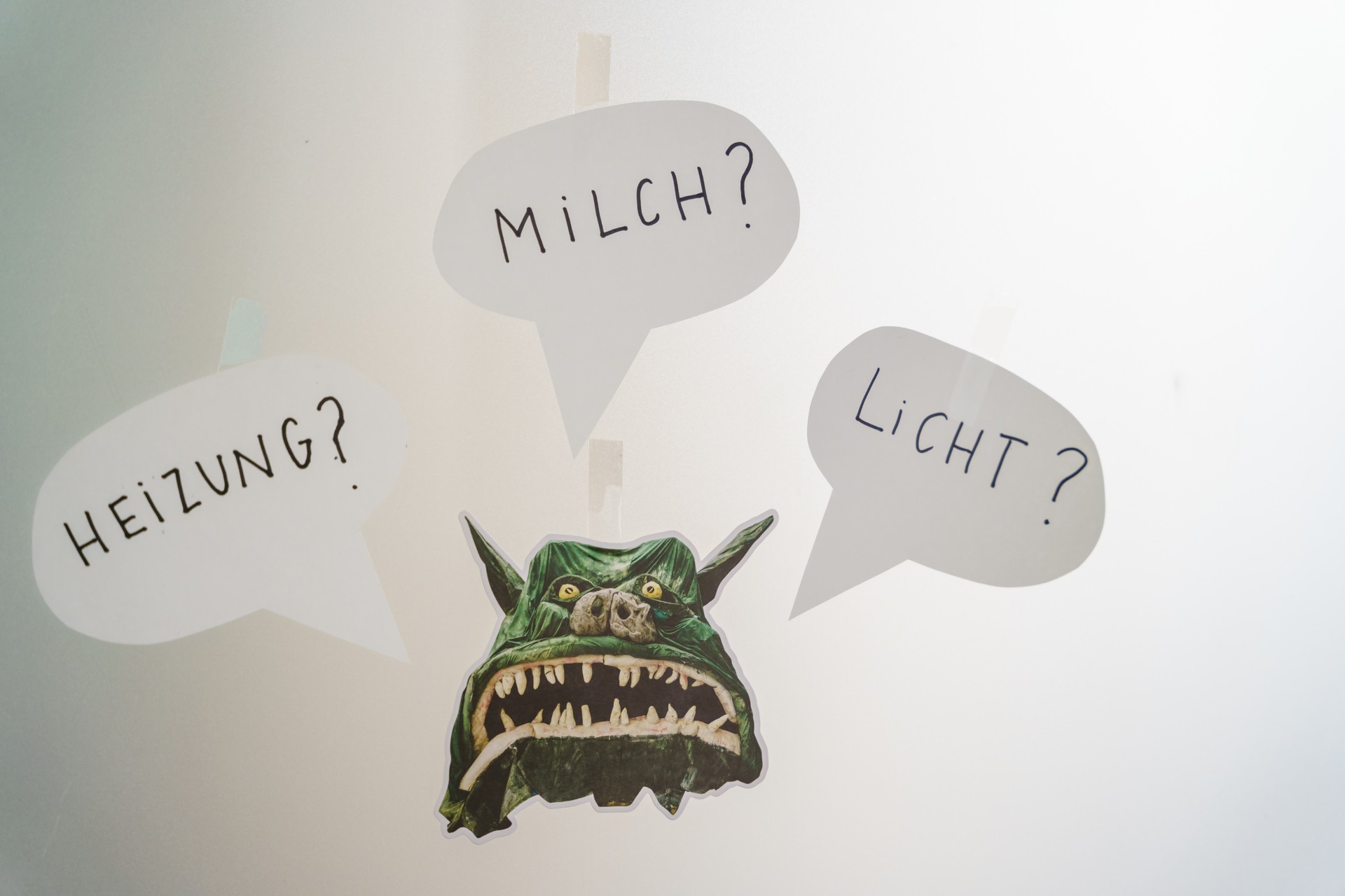 Grüner Monsterkopf mit Sprechblasen, die Fragen zu Heizung, Milch und Licht stellen.