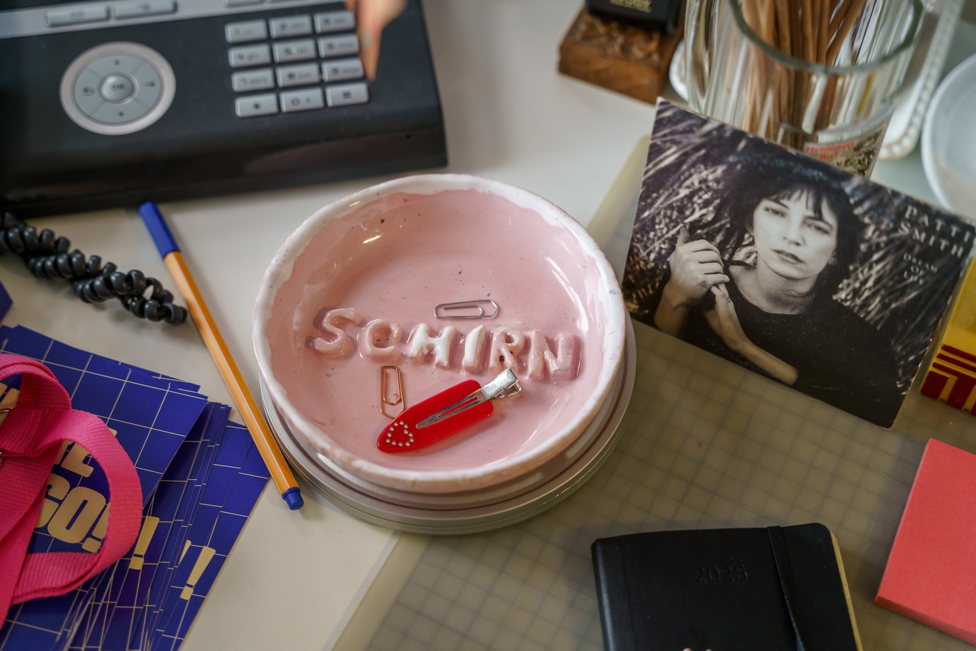 Büroplatz mit bunten Gegenständen: rosa Schale, Büroklammern, Stifte und ein Foto von Patti Smith.