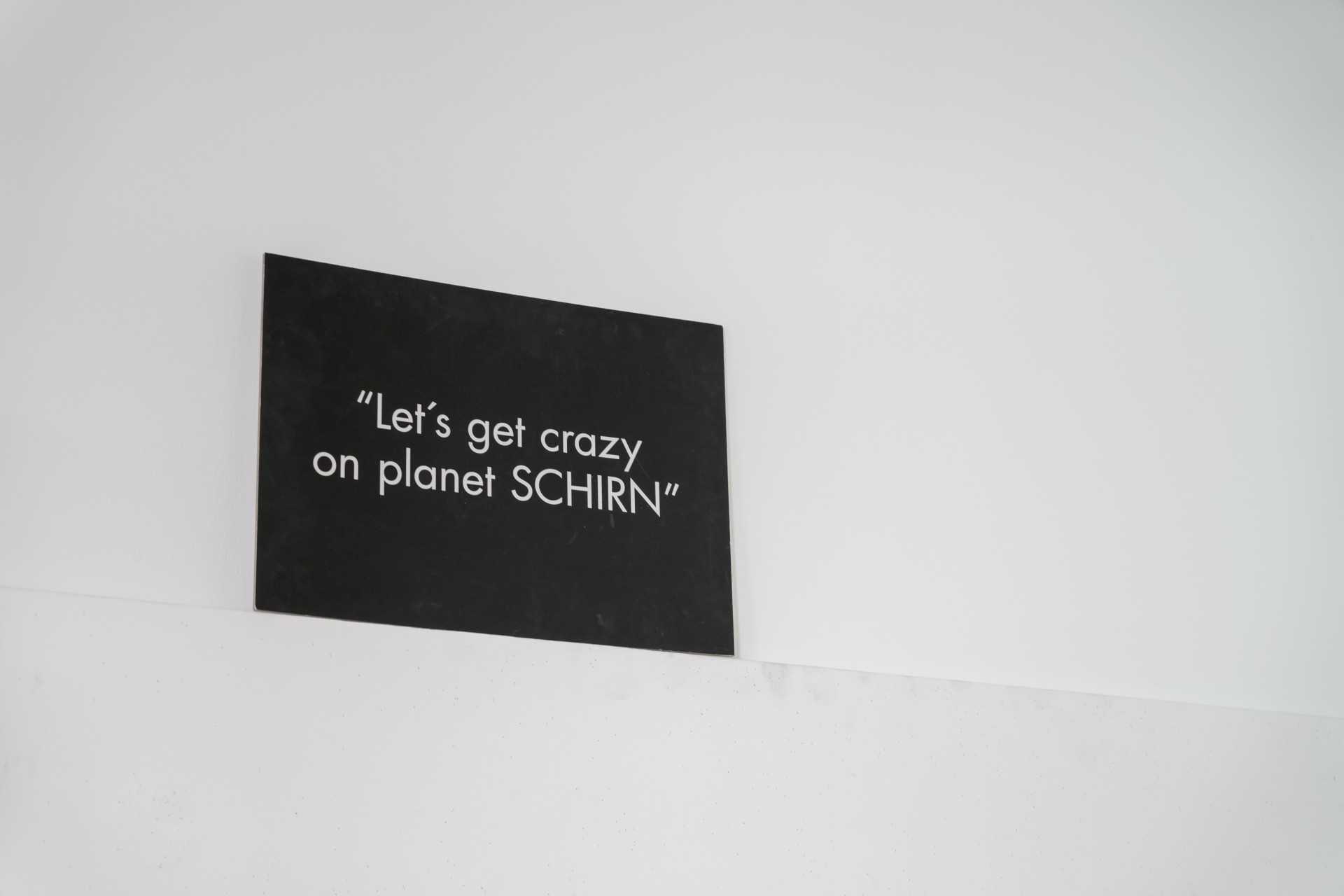 Schwarzes Schild mit dem Text: „Lass uns verrückt werden auf Planet SCHIRN“. Schlichte Hintergrundgestaltung.
