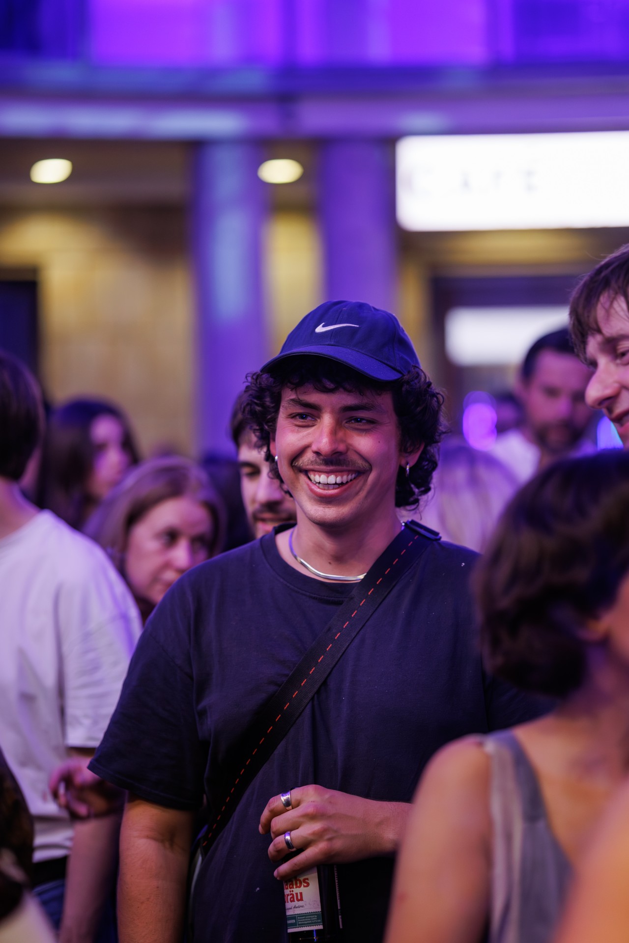 Junger Mann mit lockigem Haar und Baseballcap lächelt in einer lebhaften Menschenmenge bei einem Event.