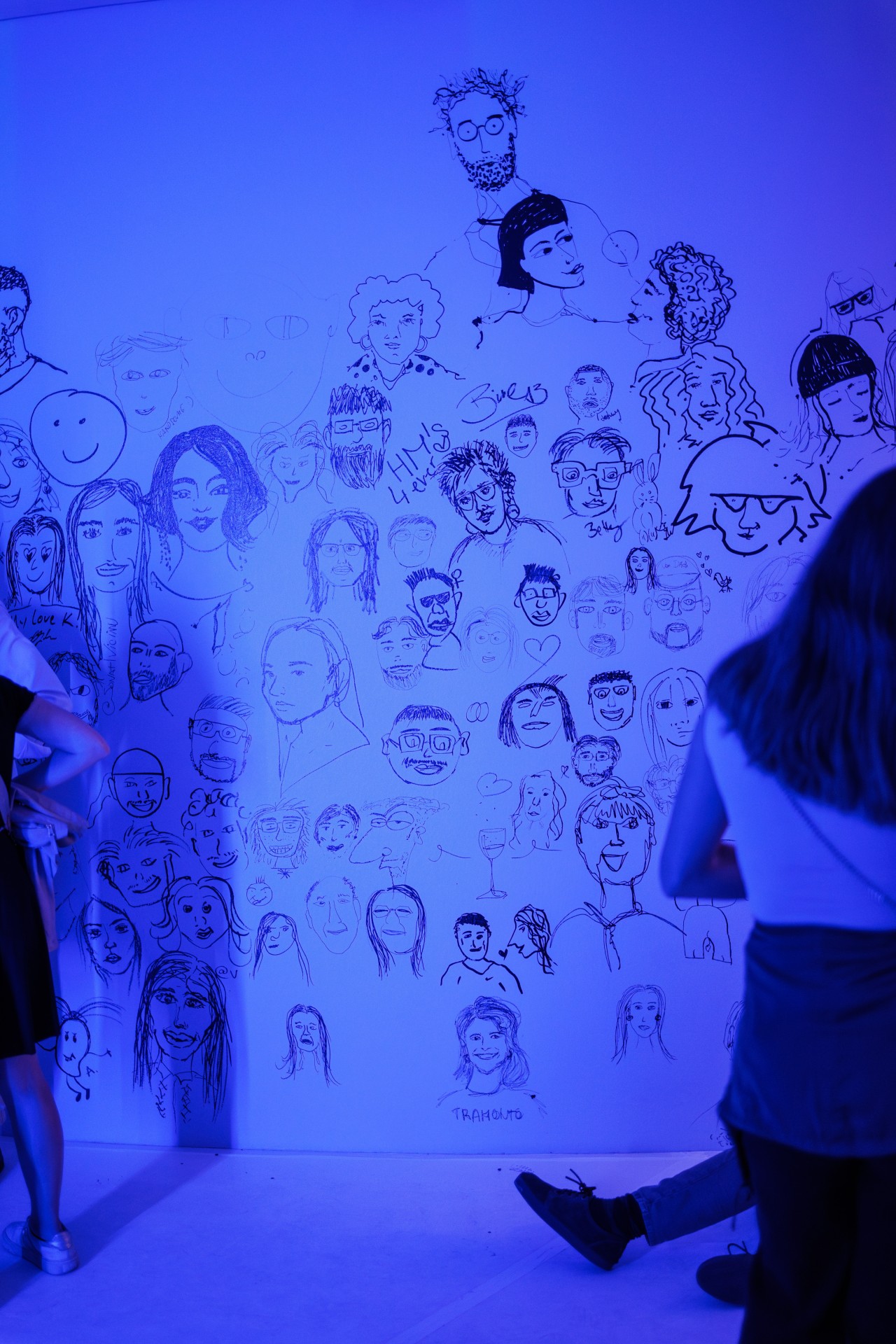 Wand mit zahlreichen Gesichtern und Figuren in blauer Beleuchtung, vor der Personen stehen.