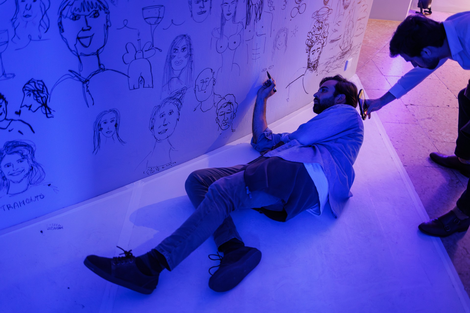 Ein Künstler skizziert Gesichter an einer Wand, während eine andere Person mit dem Smartphone filmt.
