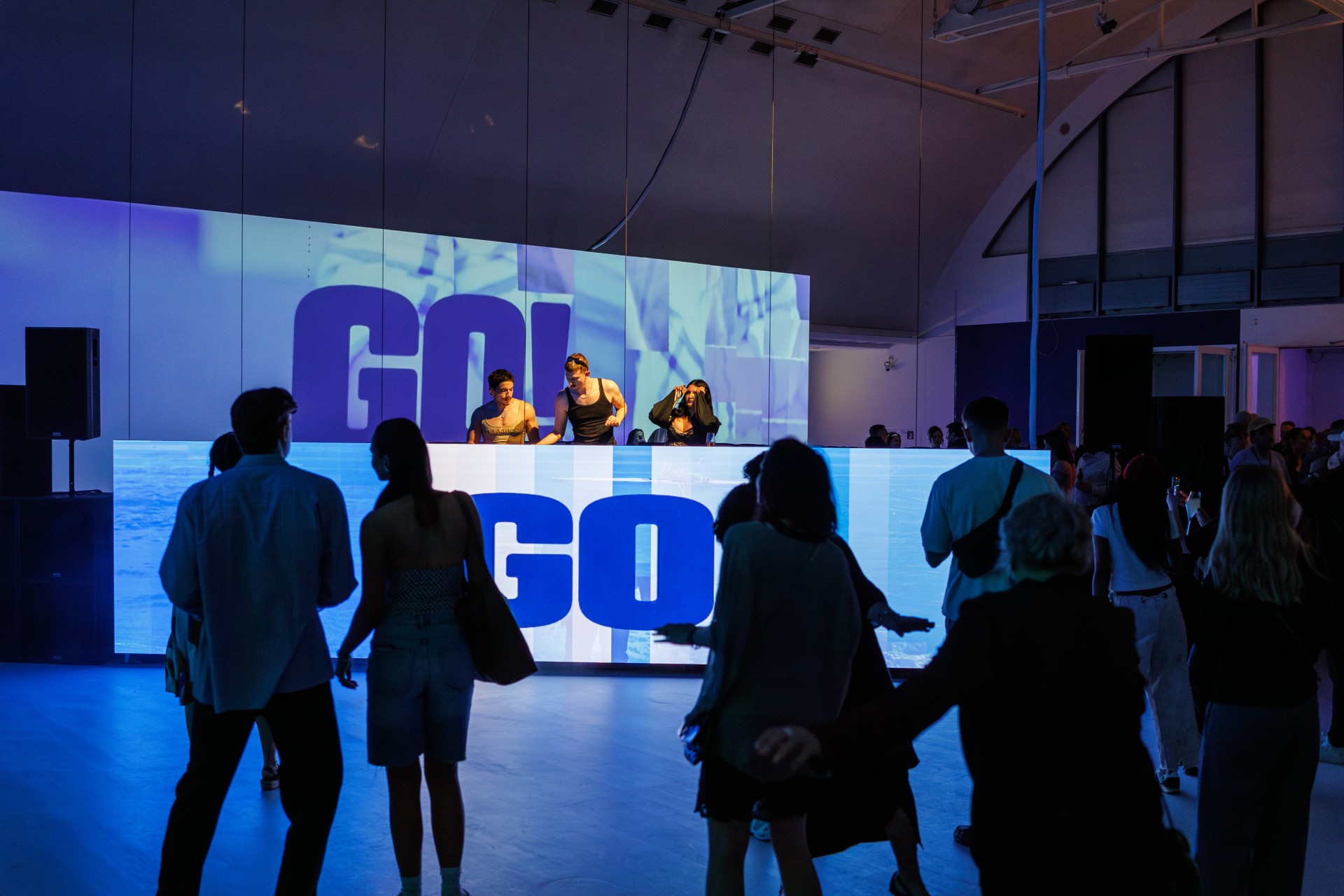 Tanzende Menschen vor DJs und einer leuchtenden "GO!"-Projektion in einem lebhaften Veranstaltungsort.