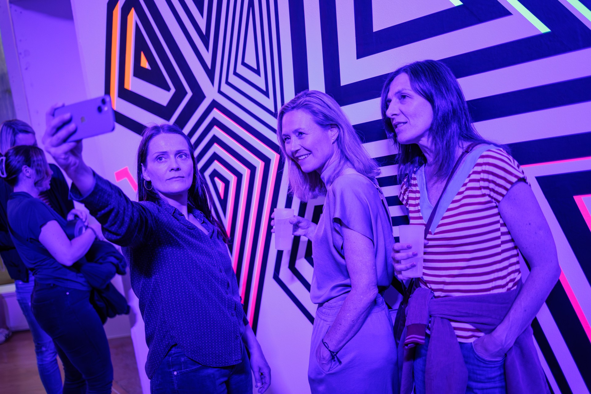 Drei Frauen posieren für ein Selfie vor einer bunten, geometrischen Wand in einem modernen Raum.