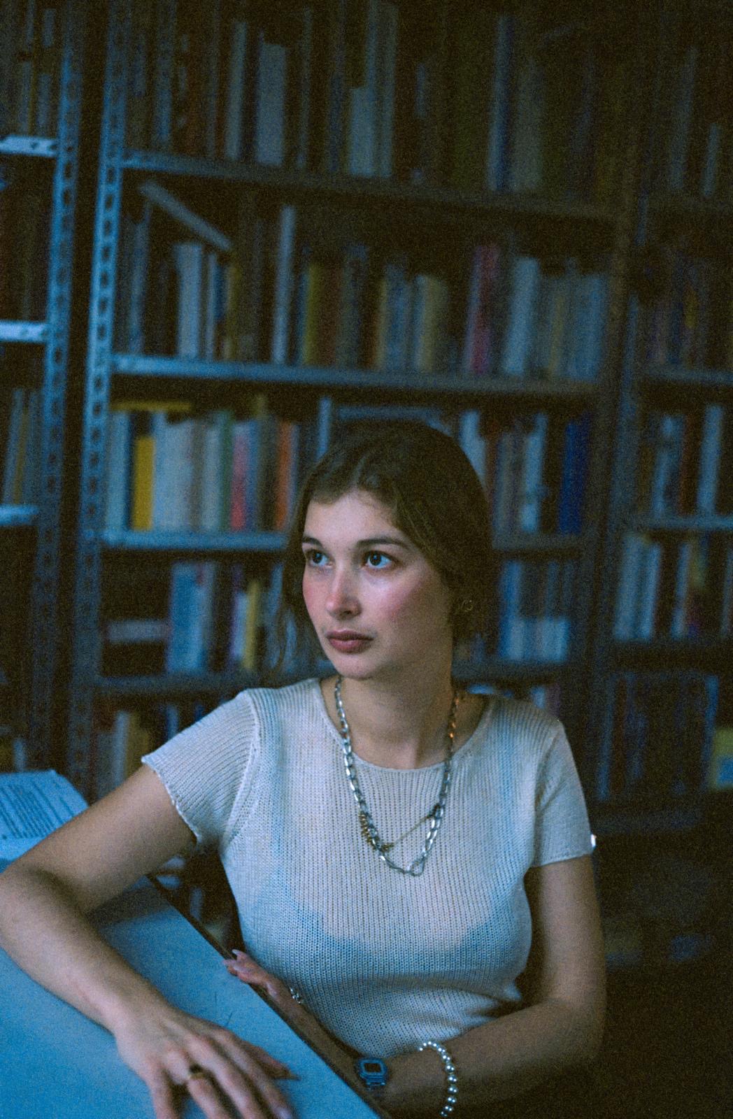 Junge Frau in einem Bücherregal, nachdenklicher Blick, trägt ein schlichtes Oberteil und Halsketten.