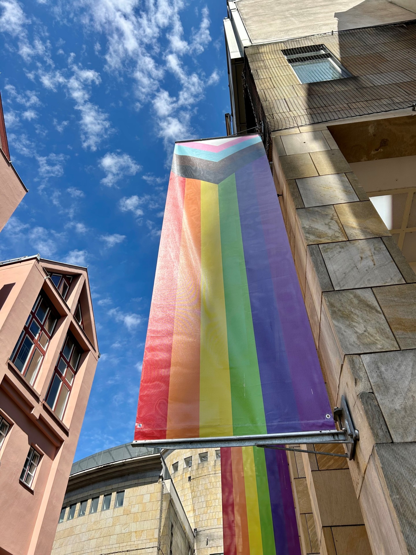 Bunte Pride-Flagge weht an einem Gebäude, blauer Himmel mit Wolken im Hintergrund.