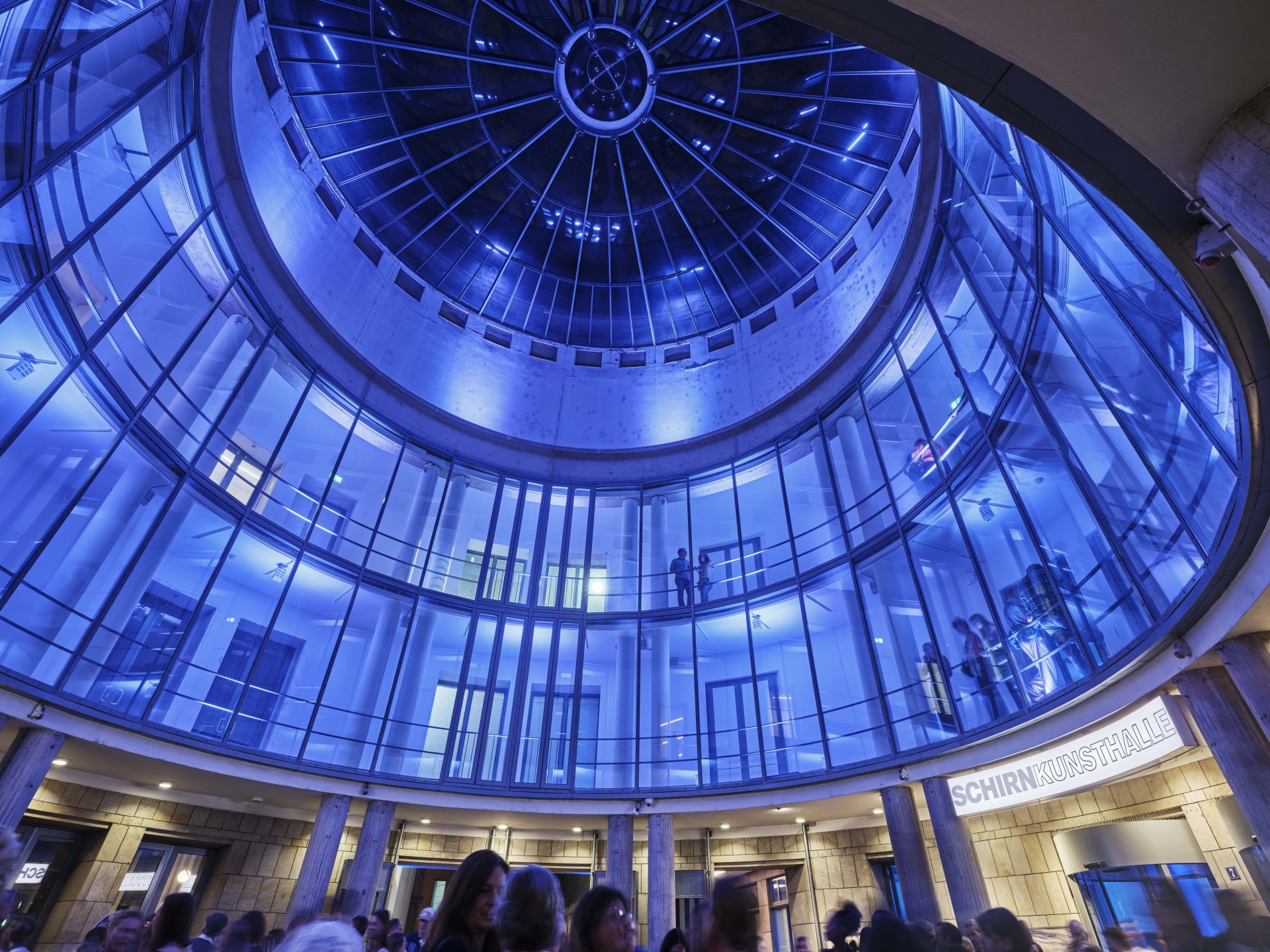 Moderne Kunsthalle mit blauer Beleuchtung, gläsernen Wänden und einer beeindruckenden Kuppel. Viele Besucher sind da.