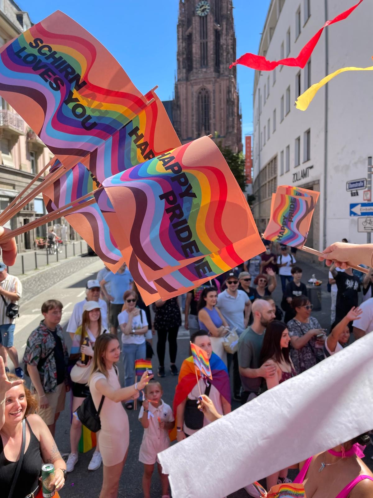 Menschen feiern Pride mit bunten Flaggen und Schildern in einer sonnigen Straße vor einem Kirchturm.