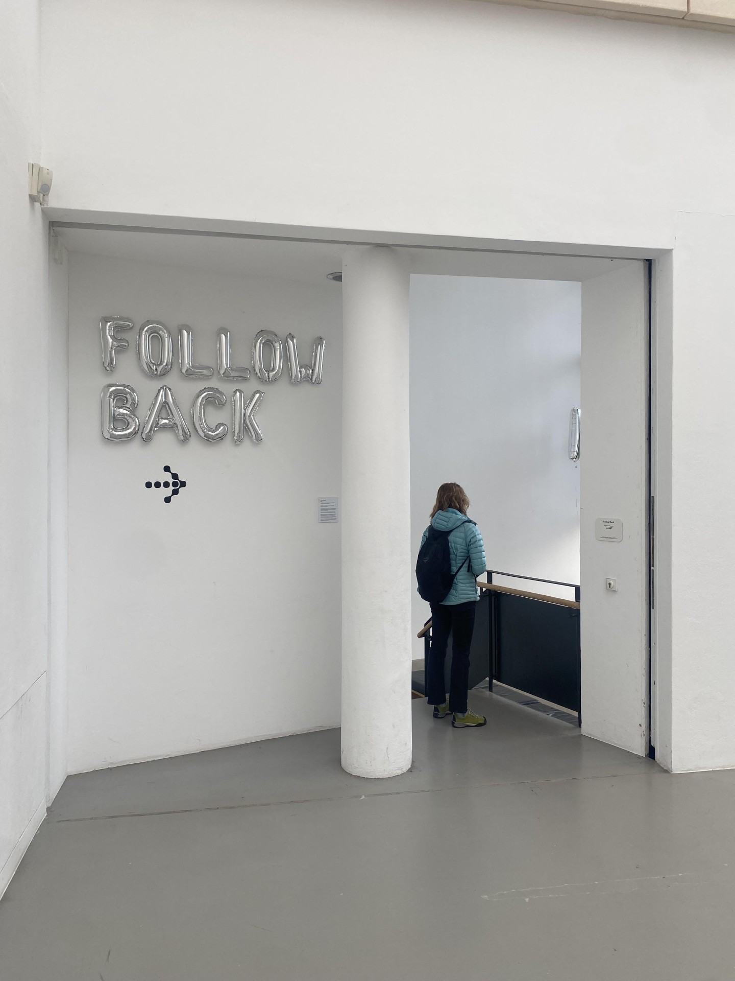 Raum mit einer Person davor; silberne Luftballonbuchstaben "FOLLOW BACK" an der Wand.