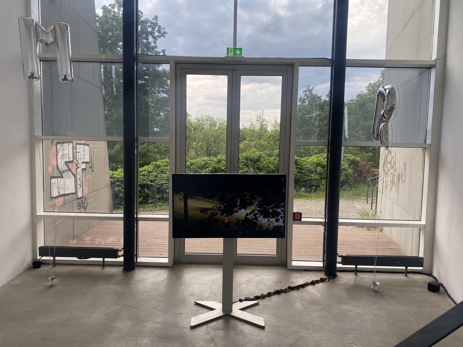 Moderne Galerie mit Fenstern, Fernseher und Luftballons. Außenbereich mit viel Grün und graffiti-verzierter Wand.