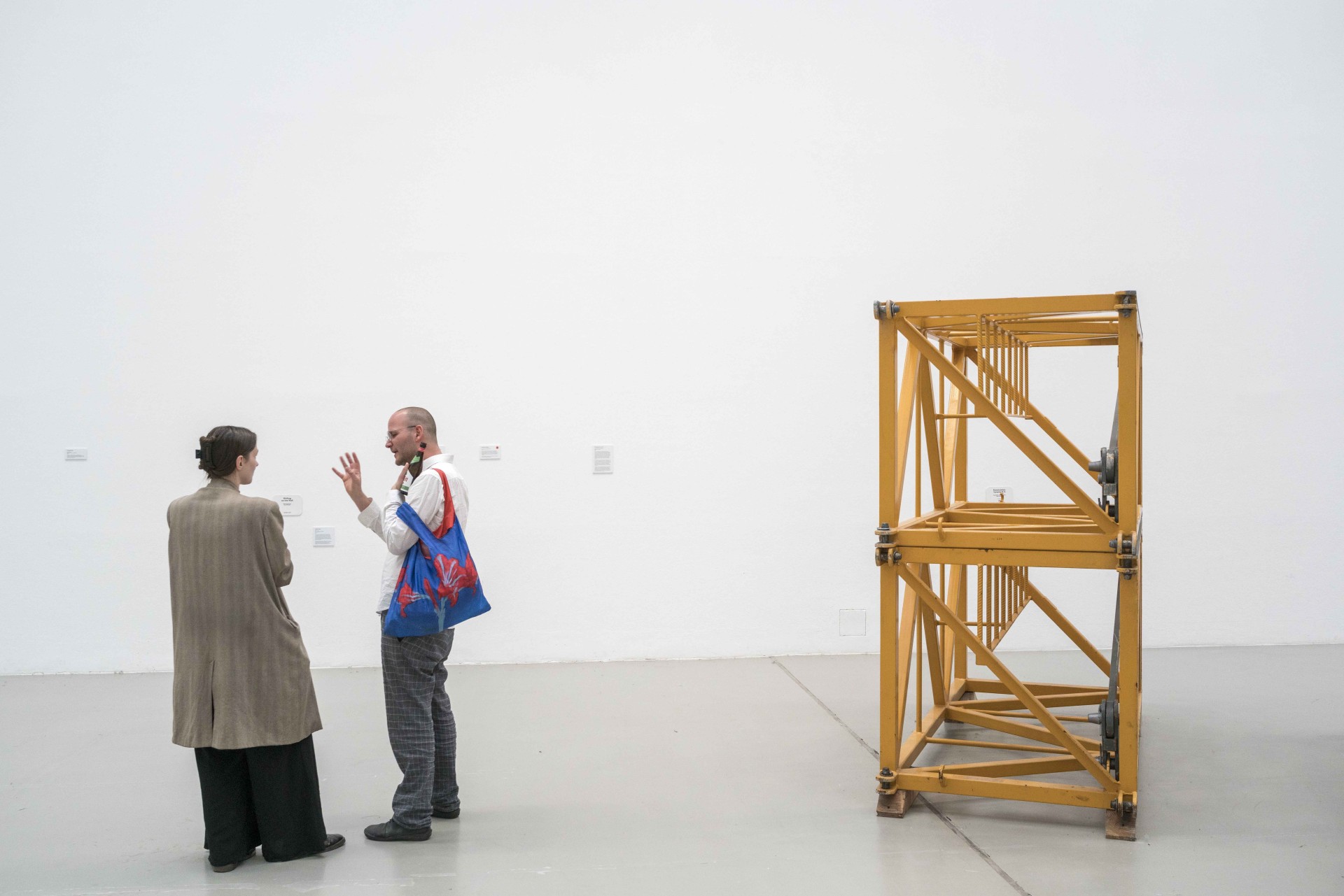 Zwei Personen unterhalten sich in einer Galerie, im Hintergrund ist ein gelbes, konstruktives Kunstwerk zu sehen.