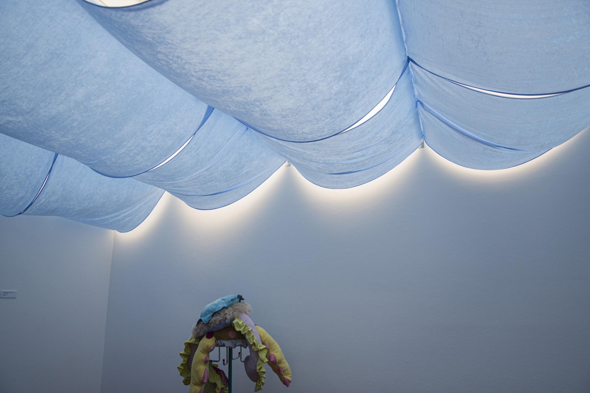 Raum mit gewellter blauer Decke und sanfter Beleuchtung; ein farbenfrohes Objekt steht im Vordergrund.
