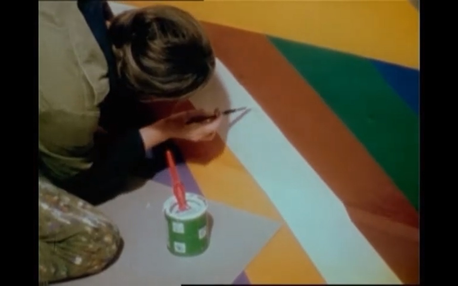 Person malt bunte geometrische Muster auf den Boden, neben einem Eimer mit Farbe und Pinsel.