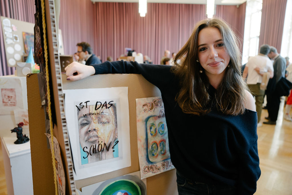 Junge Frau lächelt vor Kunstwerken, eines fragt: "Ist das wirklich schön?" Im Hintergrund sind Besucher und Kunst zu sehen.