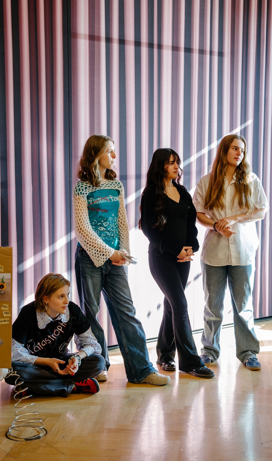 Vier Jugendliche stehen vor einer gestreiften Wand. Eine sitzt, die anderen stehen in verschiedenen Outfits.