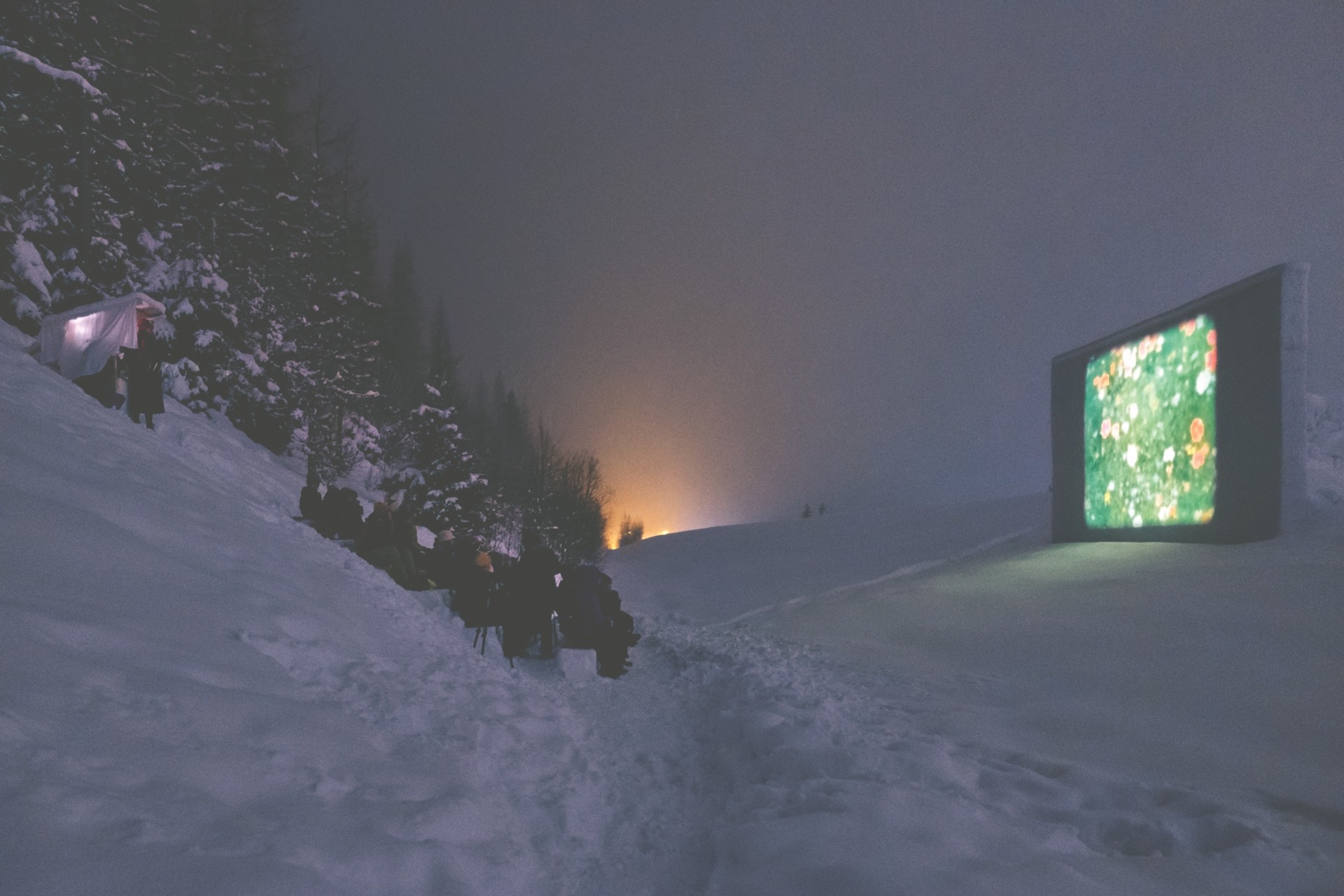 Outdoor-Kino auf schneebedecktem Gelände mit Zuschauern, einem Projektor und nebligem Hintergrund.