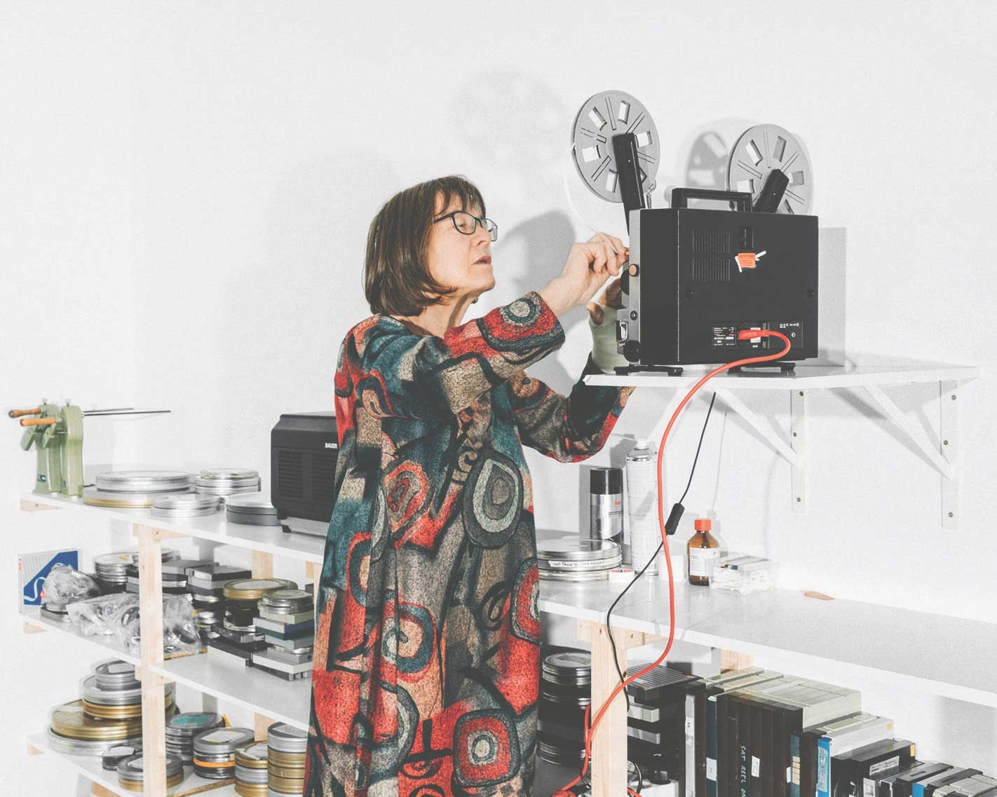 Eine Frau in einem bunten Kleid arbeitet an einem Filmprojektor, umgeben von Filmrollen und Ausrüstung.