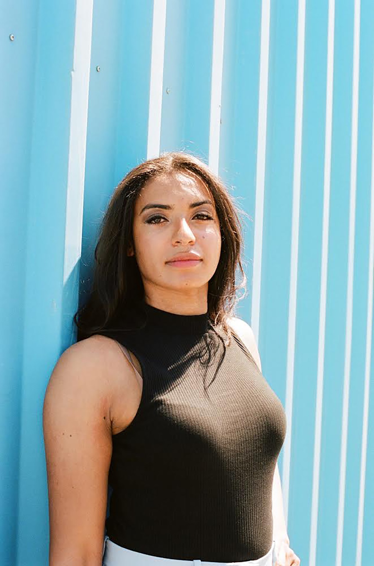 Young woman in a black top leans against a blue wall, light highlighting her friendly face and natural beauty.