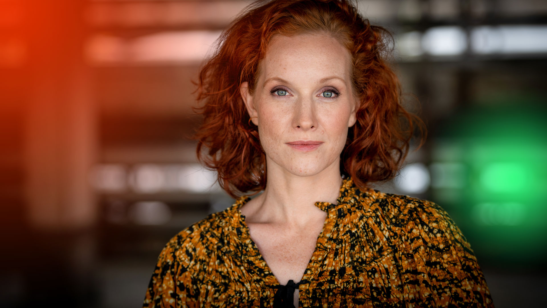 Porträt einer Frau mit roten Locken in buntem Oberteil, vor einem modernen, unscharfen Hintergrund aufgenommen.