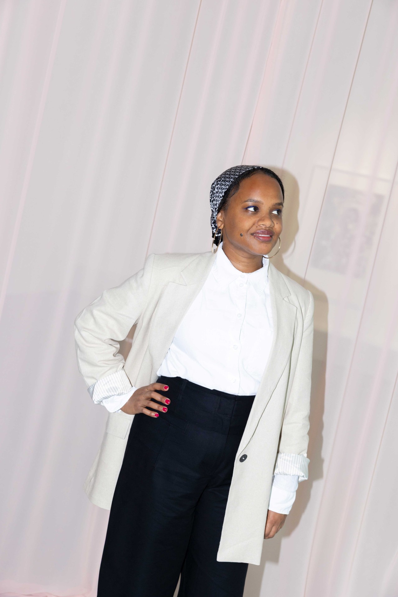 A woman in elegant attire with a pale blazer and dark pants, smiling against a soft pink background.