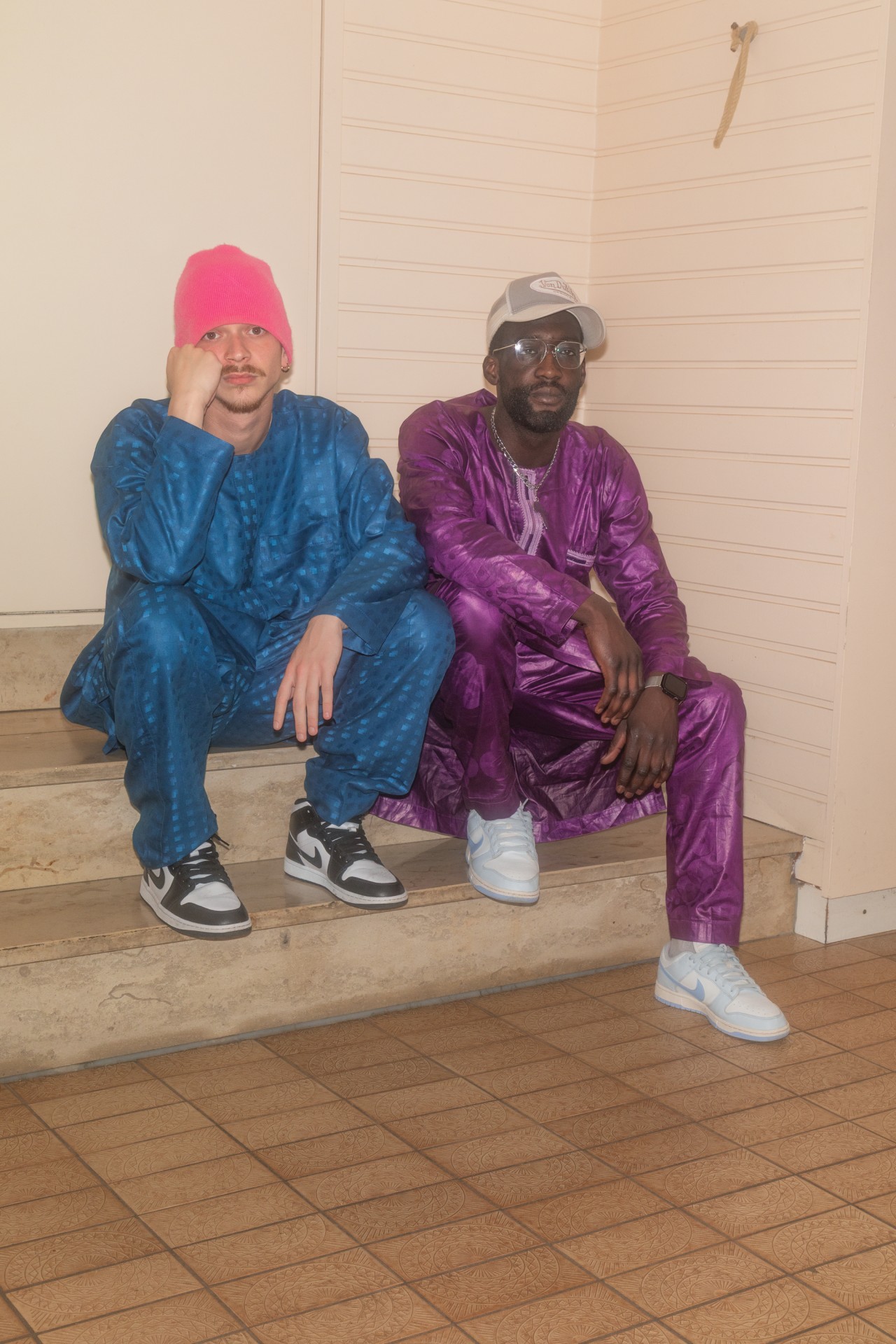 Zwei Männer sitzen auf einer Treppe: Einer trägt einen pinken Hut und blauen Anzug, der andere einen lila Anzug und eine Kappe.