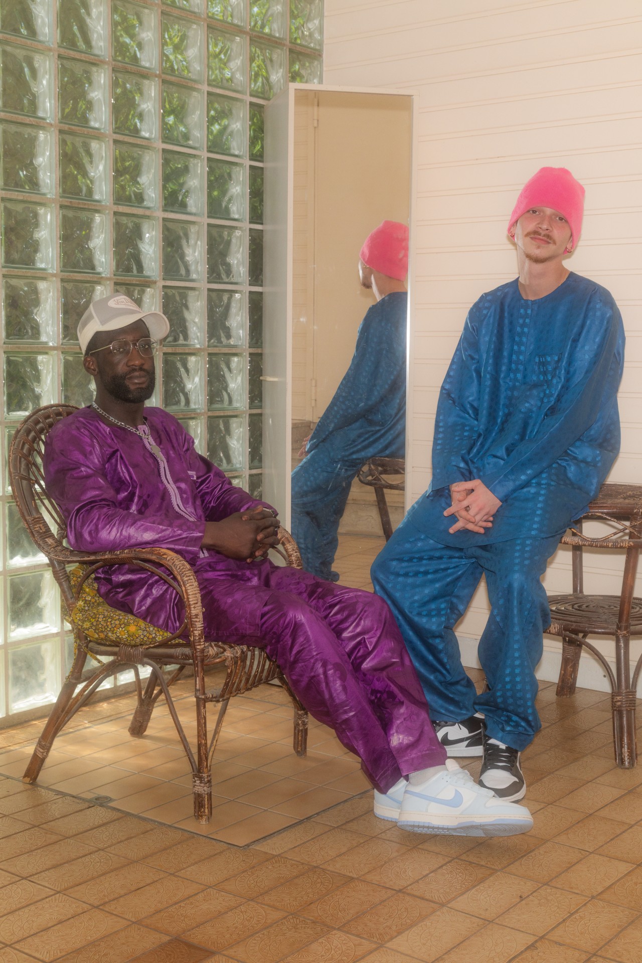 Zwei Männer in bunten Outfits und Sneakers sitzen in einem hellen Raum mit großen Fenstern und einem Spiegel.