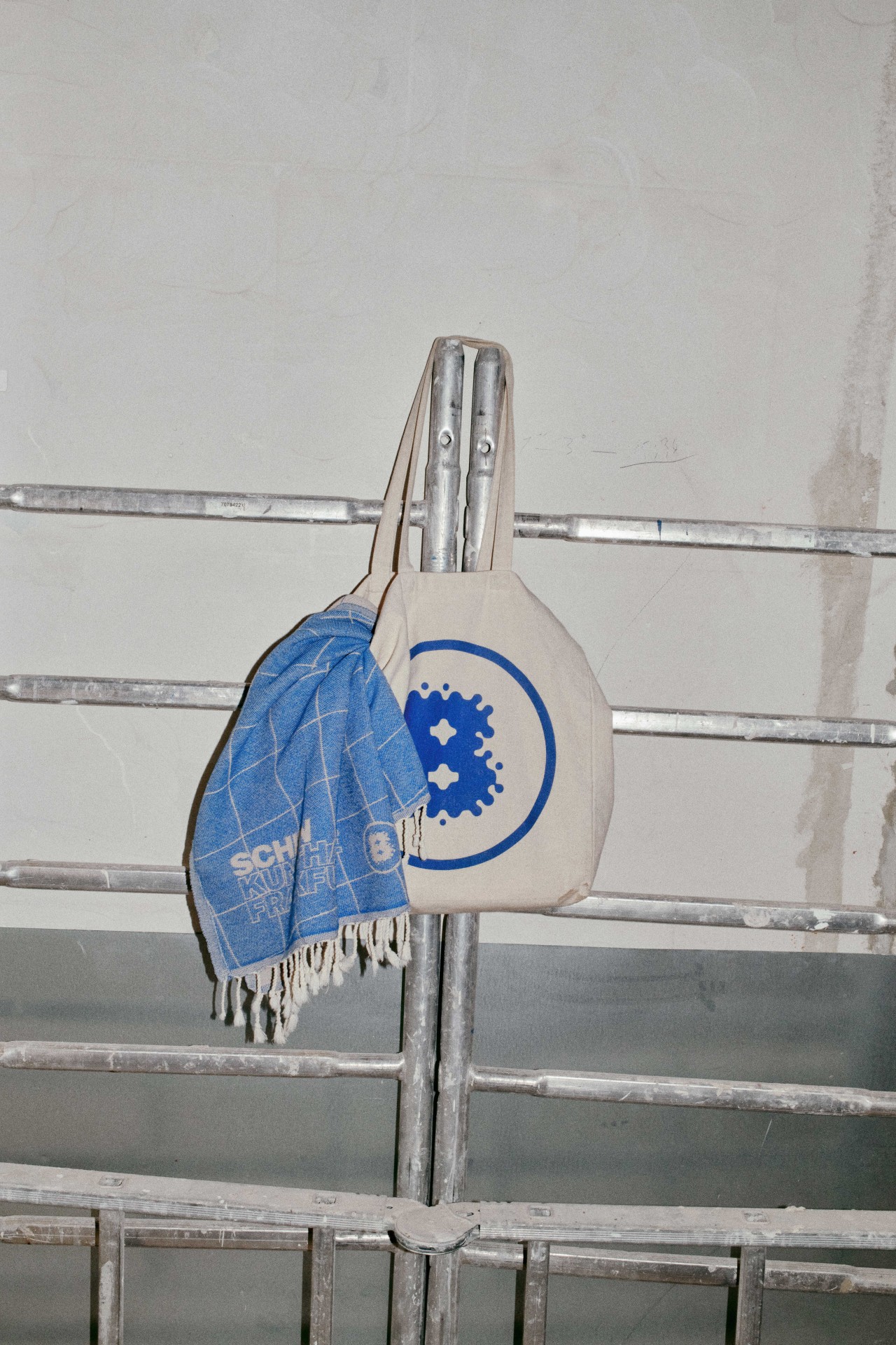 Beige Tasche mit blauem Logo und blauer Decke hängen an einem Metallgestell in einem hellen Raum.