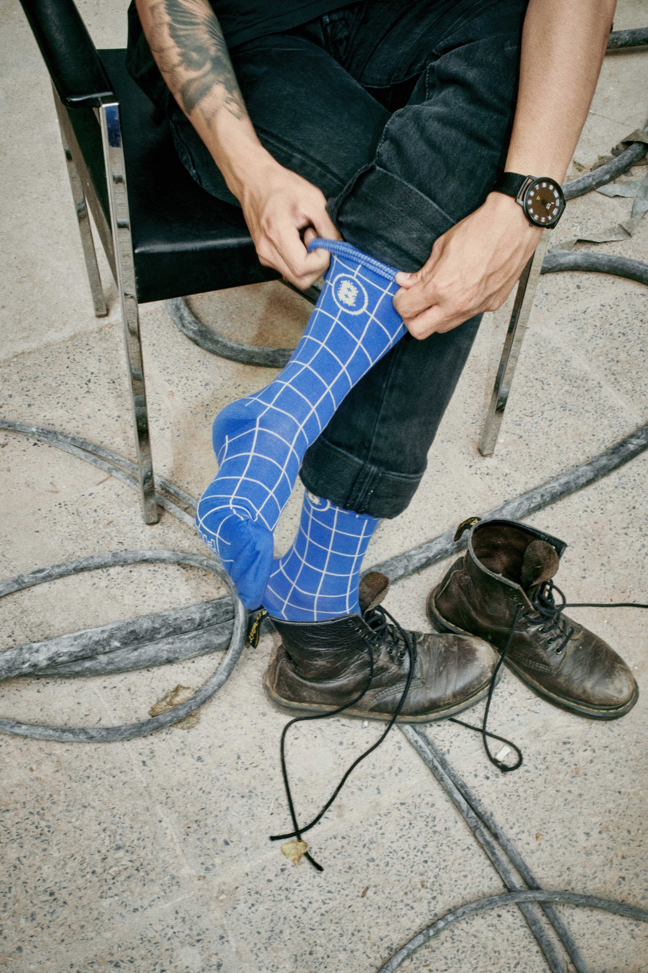 Mann sitzt auf einem Stuhl, trägt blaue Karosocke und Boots und passt seine Socke an.