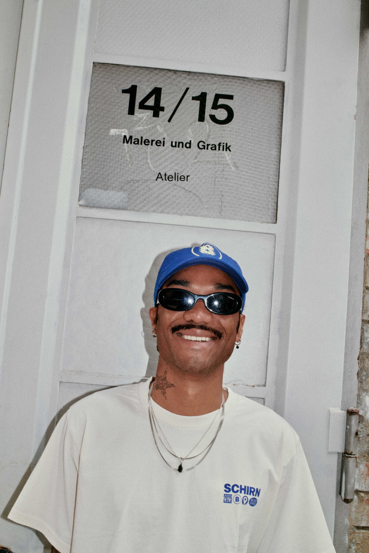 Junger Mann mit Sonnenbrille und Kappe, lächelnd vor Atelier-Tür mit der Aufschrift „14/15 Malerei und Grafik“.