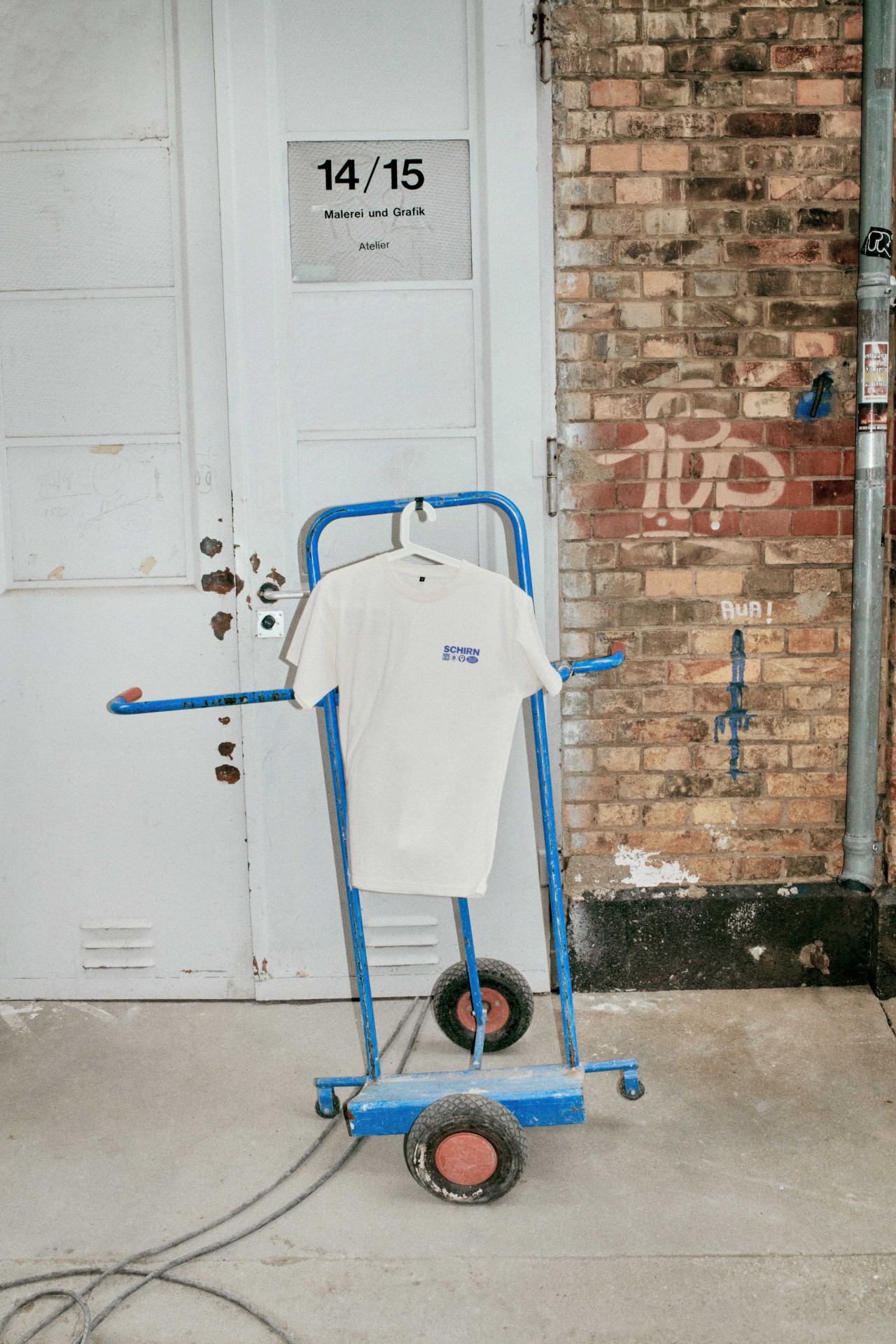 Ein blauer Wagen mit einem weißen T-Shirt steht vor einer grauen Tür mit der Aufschrift "14/15 Malerei und Grafik".