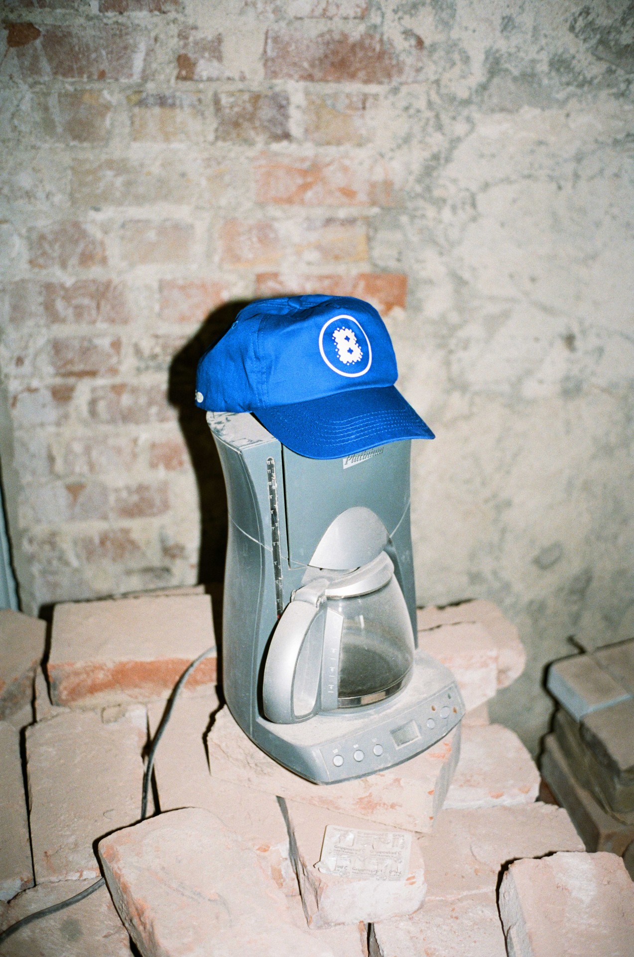 Eine blaue Cap liegt auf einem Kaffeekocher, der auf einem Stapel Ziegelsteine vor einer Backsteinwand steht.