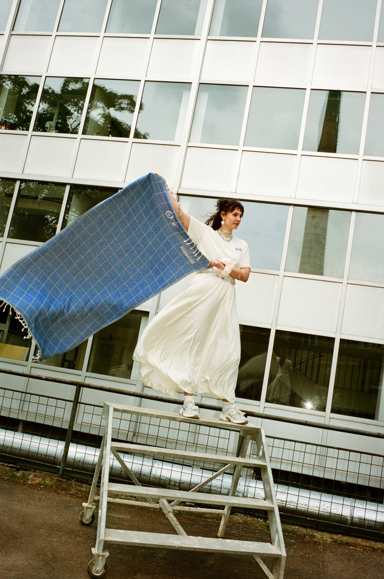 Frau im weißen Kleid steht auf einem Gerüst und hält eine blaue Decke vor einem modernen Gebäude.