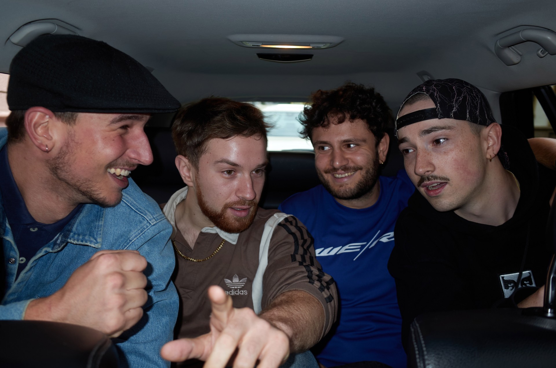 Vier Männer lachen und unterhalten sich im Auto, jeder trägt ein anderes Outfit.