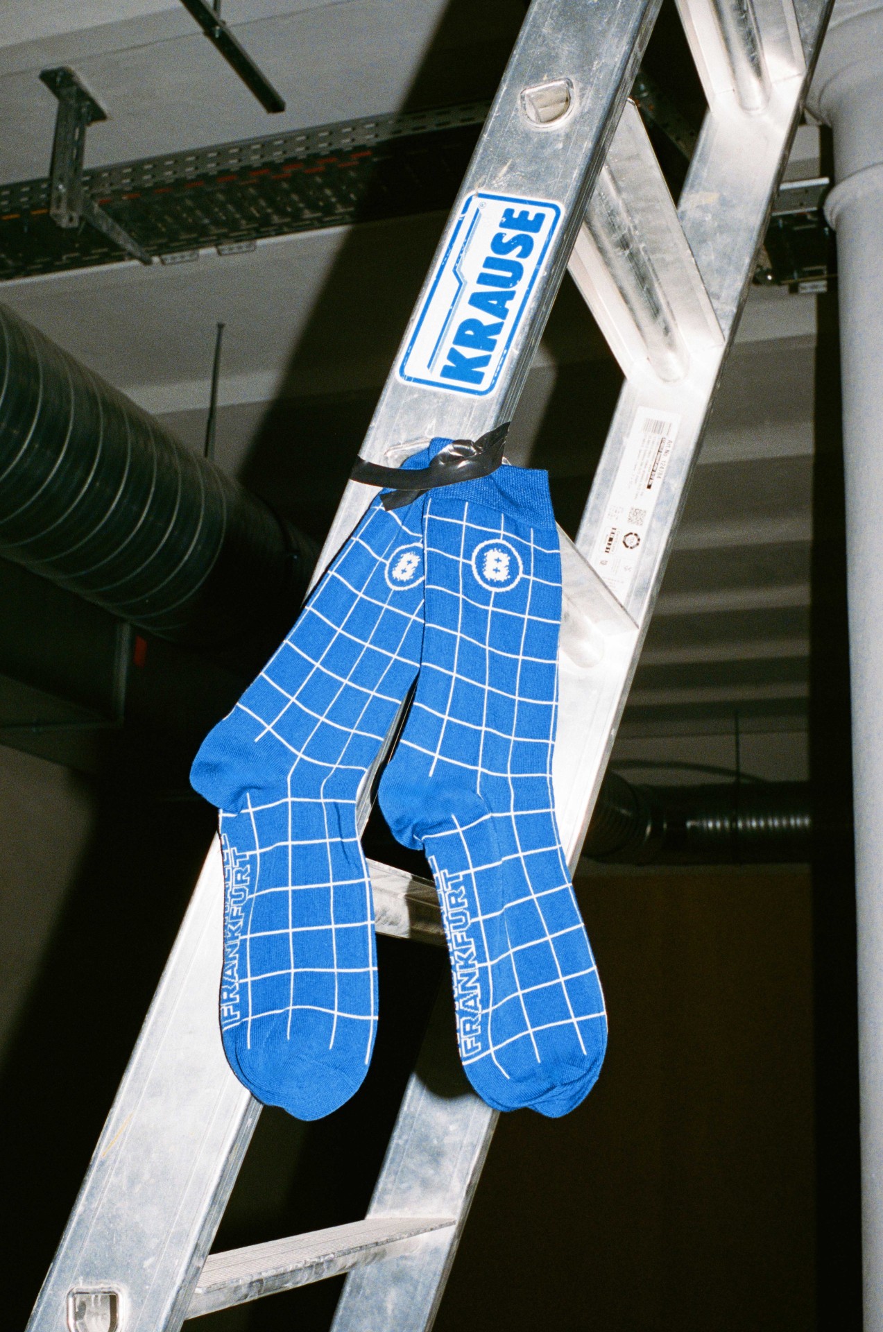 Blaue Socken mit quadratischem Muster hängen an einer Aluminiumleiter in einem industriellen Raum.