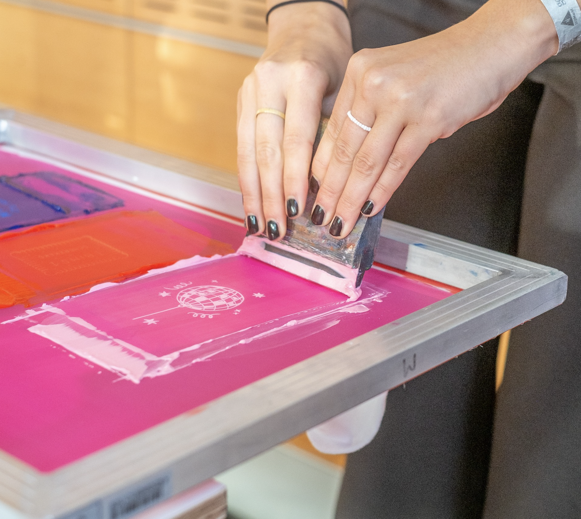 Hände mit dunklem Nagellack ziehen einen Rakel über einen pinken Siebdruckrahmen, sichtbar sind die Farbschichten.