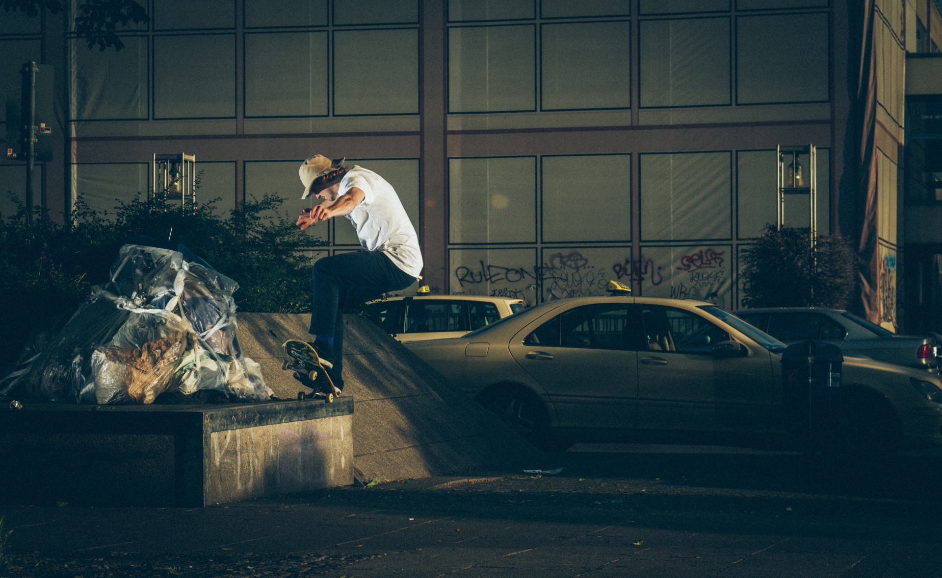 Skateboarder springt über Müll auf einer Bank, im Hintergrund Autos und ein mit Graffiti besprühtes Gebäude.