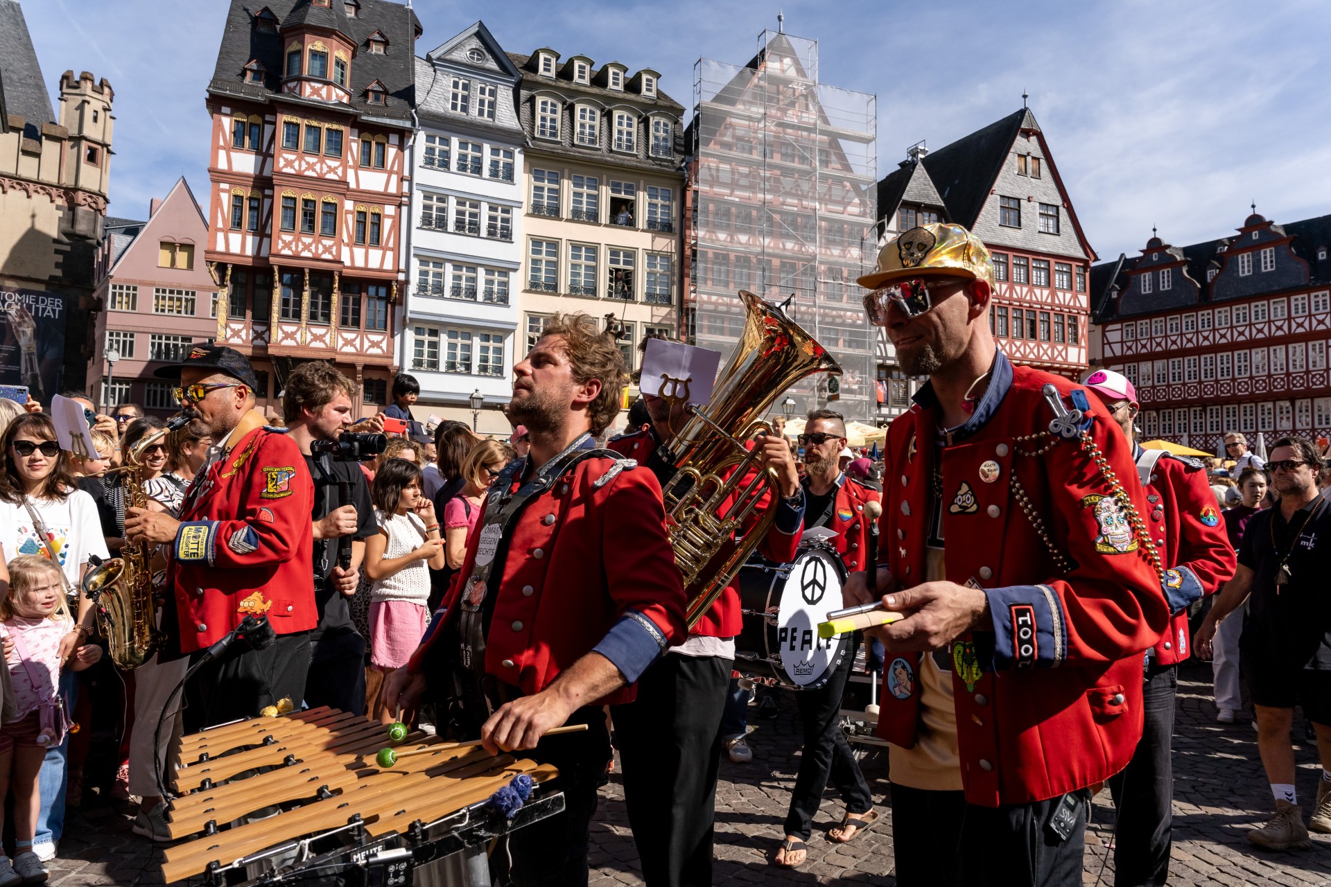 Lebhafte Parade mit Musikern in roten Jacken auf einem Platz, umgeben von fröhlichen Zuschauern und historischen Gebäuden.