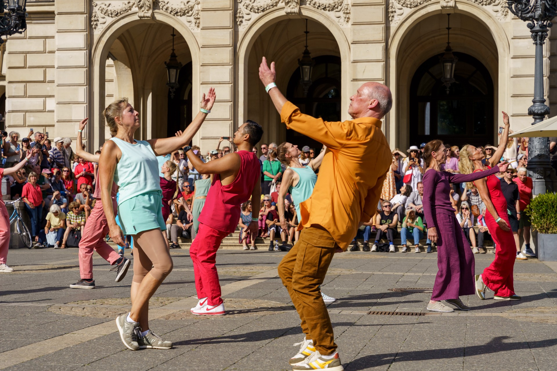 Tanzende Menschen in bunten Outfits vor einem Publikum, das eine Aufführung auf einem Platz verfolgt.