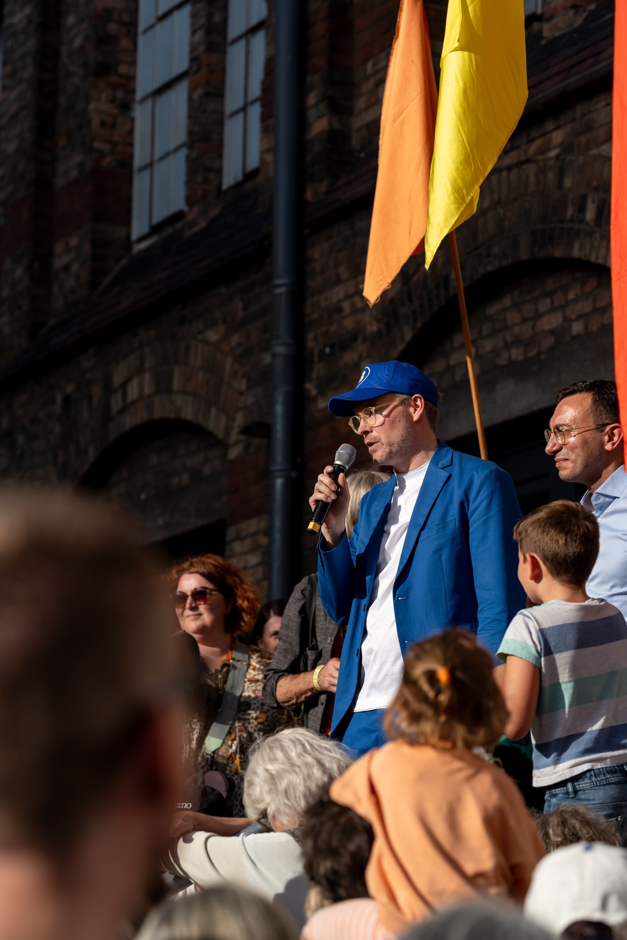 Mann spricht mit Mikrofon auf Bühne, umgeben von einer Menschenmenge mit bunten Fahnen.