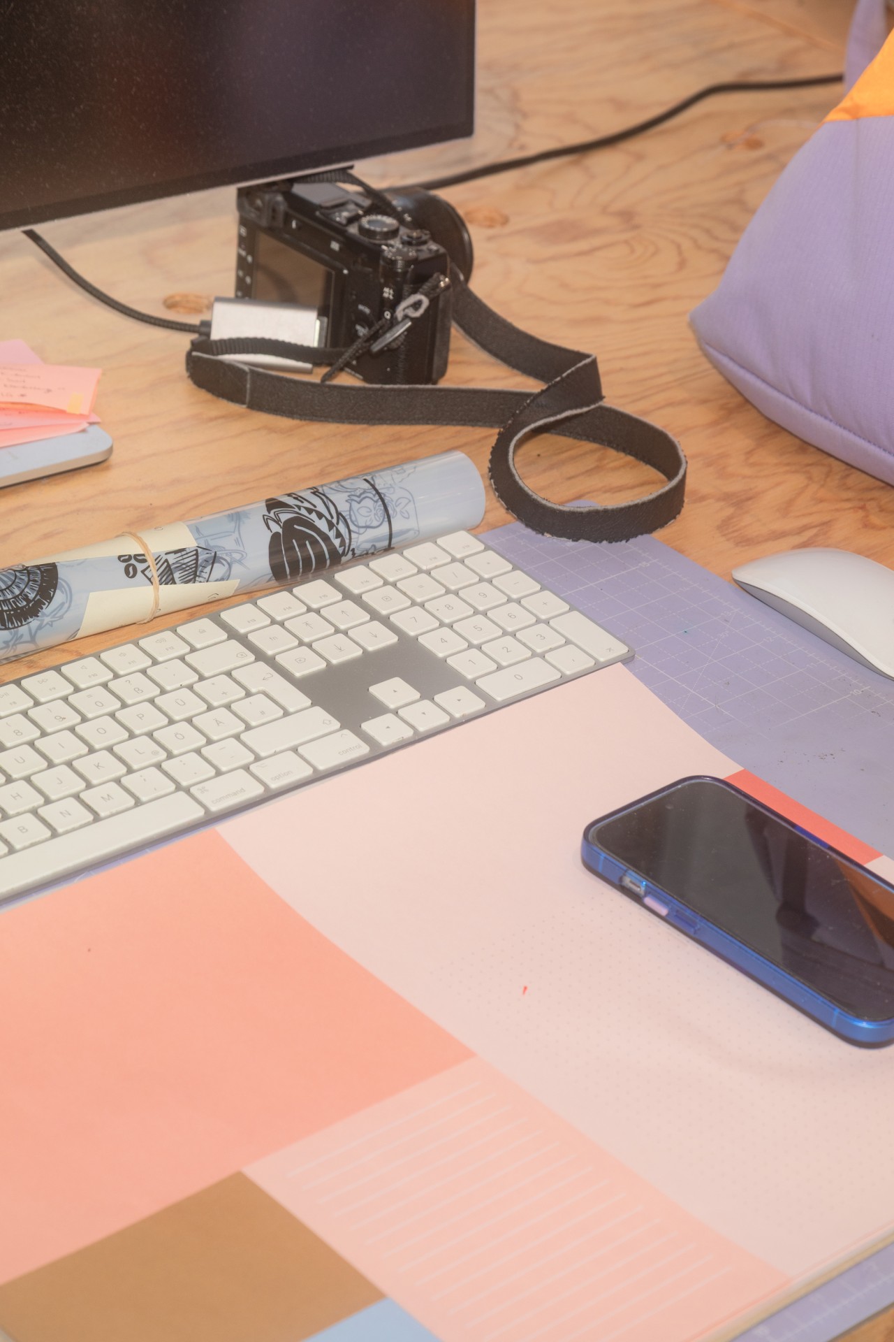 Arbeitsplatz mit Computer, Kamera, Smartphone und buntem Hintergrund. Notizen und eine gezeichnete Papierrolle sichtbar.